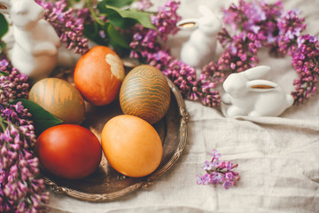 Happy Easter. Stylish Easter eggs on rustic plate, white bunnies and lilac flowers on linen rural fabric. Top view. Natural dyed yellow easter eggs and spring flowers. Holiday decor.