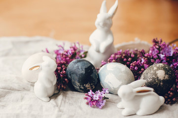 Happy Easter. Modern Easter eggs, white bunnies and lilac flowers on linen rustic fabric. Stylish holiday table decor. Space for text. Natural dyed easter eggs and spring flowers.