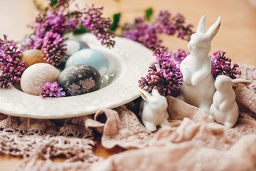 Stylish Easter eggs on vintage plate, white bunnies and lilac flowers on fabric on wooden table. Rural composition of colorful natural dyed easter eggs and spring flowers. Holiday decor
