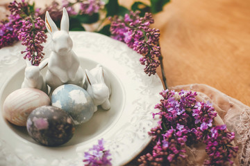 Obraz na płótnie Canvas Happy Easter. Stylish Easter eggs on vintage plate, white bunnies and lilac flowers on fabric on wooden table. Rural composition of colorful natural dyed easter eggs and spring flowers