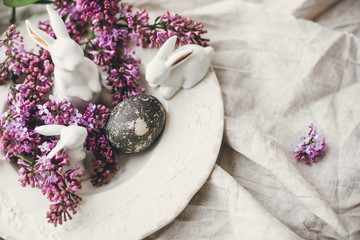 Happy Easter. Stylish Easter white bunnies, modern easter egg on vintage plate and lilac flowers on linen rural fabric. Natural dyed easter egg and spring flowers. Space for text