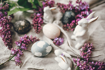 Obraz na płótnie Canvas Modern Easter eggs, white bunnies and lilac flowers on linen rural fabric. Stylish holiday table decor. Happy Easter. Space for text. Natural dyed easter eggs and spring flowers.