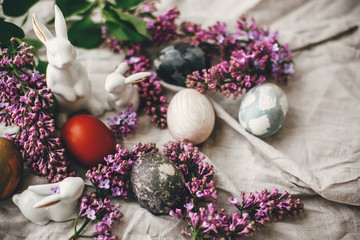 Obraz na płótnie Canvas Happy Easter. Modern Easter eggs, white bunnies and lilac flowers on linen rural fabric. Stylish holiday table decor. Space for text. Natural dyed easter eggs and spring flowers.