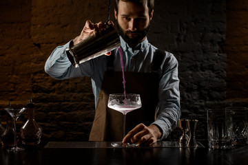 Professional bartender pouring a violet alcoholic drink to the big cocktail glass from the steel...