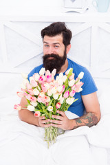 Make surprise concept. Gift for spouse. Bearded hipster in bed. Spring in bedroom. Man hold tulips bouquet while relaxing in bed. Fresh flowers. Flowers delivery service. Birthday anniversary holiday