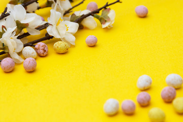 Branch of white flowers on yellow background with candy, easter chocolate eggs. Easter mock up. Minimalistic Spring background.