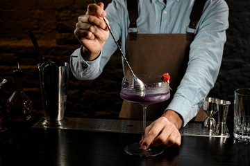 Male bartender stirring a violet alcoholic drink in the big cocktail glass decorated with a red flower