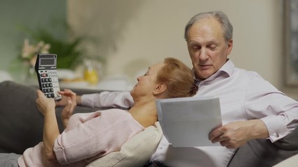 Loving senior couple calculating home finances together. Man sitting on sofa and reading data on paper bills, wife lying on his lap and using calculator - Powered by Adobe
