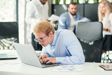 Cheerful purposeful male office worker with funny eyeglasses sits at his workplace and using laptop to solve financial tasks.