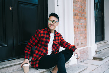 Cheerful ethnic casual guy sitting on doorstep while drinking takeaway coffee