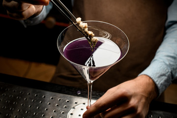 Professional male bartender decorated purple alcoholic cocktail in a martini glass with a flower by tweezers