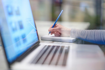 Closeup of hand woman writing working with a pen on a document, close to her computer laptop pc in her office on her workplace, desktop career screen notebook business, cold blue atmosphere blurred