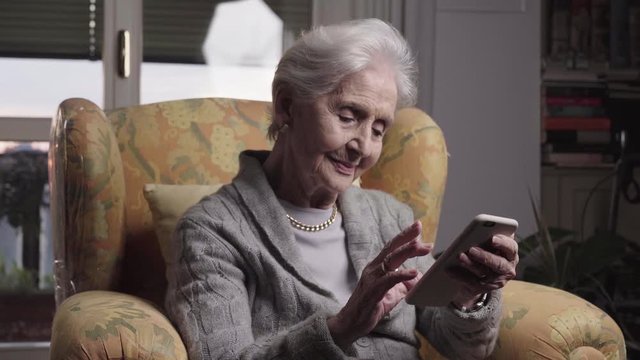 94 years old skilled lady portrait using smartphone technologies to communicate easily from her living room, senior woman sitting in yellow armchair holding phones and texting to family. 