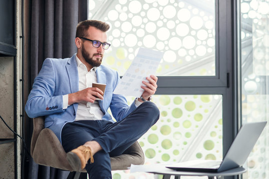 Close up of industrious good-looking young businessman analising document in his hand and drinking fragrant coffee in specially designated room in modern office