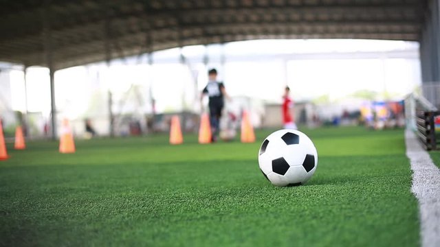 Football on green artificial turf with blurry kid soccer team training. Blurry kid soccer player running and control ball between cone markers with coach and soccer equipment in soccer academy.