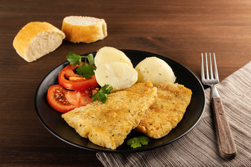 Closeup of a delicious baked battered fish seasoned with fresh parsley and accompanied by cooked potatoes, fresh tomato and freshly baked bread. On a rustic wooden table. Space for text.