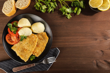 Top view of a food background with a delicious baked battered fish with fresh parsley, lemon, cooked potatoes, fresh tomato and freshly baked bread. Space for text. On a rustic wooden table.