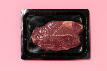 Black plastic pack with fresh beef steak isolated on pink background. Raw meat packed without label...