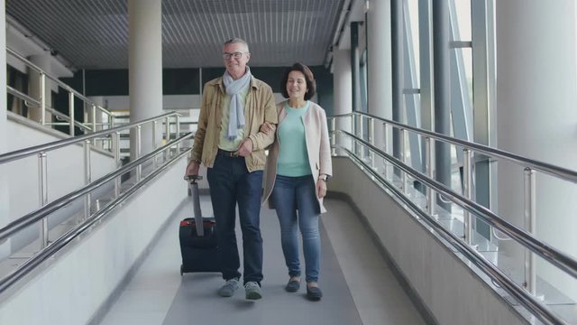 Senior couple walking with luggage in airport walkway. Aged travelers going to board plane