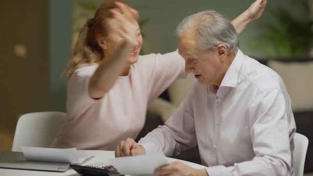 Happy elderly couple calculating home finances together sitting at table at home, looking at paper bills and using calculator. Cheerful wife embracing husband being happy with high revenue