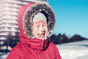 Fototapeta na wymiar Funny cute Caucasian smiling girl in red jacket parka squinting eyes from bright sun. Happy kid with snow on face at cold winter sunny day. Winter seasonal outdoor fun activity for children.