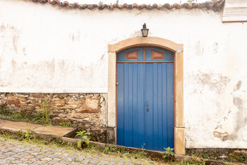 Typical colourful doorway facade on steep cobble street in Ouro Preto, Minas Gerais, with a blue door in a white wall elegantly framed by natural stone doorpost and light above