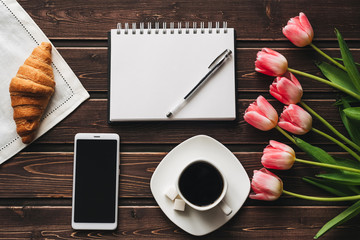Cup of coffee with a croissant for Breakfast on the table decorated with a bouquet of pink tulips and a smartphone