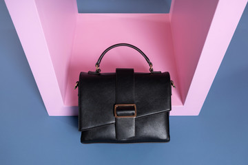 a small female black leather rectangular handbag with a handle stands on a pink, blue stand of an interesting shape. Studio photo