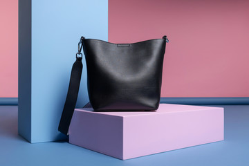 a women's black leather rectangular handbag with a shoulder strap stands on a pink, blue stand in an interesting shape. Studio photo