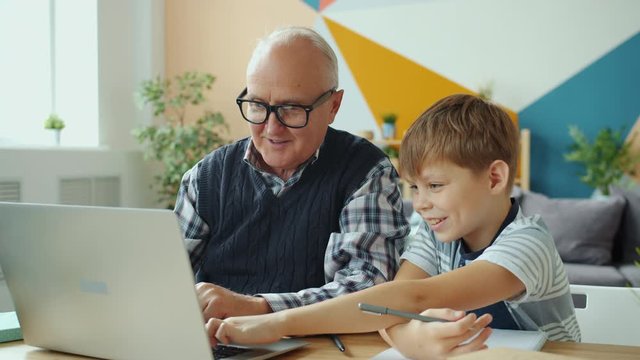 Retired grandfather and happy grandson are using laptop talking laughing working at desk in house. Contemporary lifestyle, computing and family concept.
