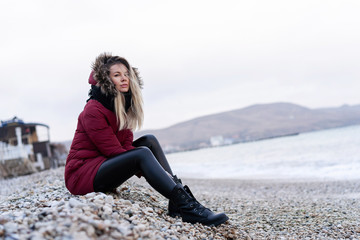 young beautiful caucasian girl in a red warm jacket with a hood, scarf, leather tight pants sitting on a rocky beach resting after a walk by the sea © Денис Бухлаев
