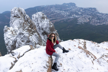 beautiful caucasian girl in a red park, warm jacket, jeans, black leather boots and sunglasses posing on top of a snowy mountain. Beautiful view