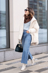 European model girl in a beige oversized down jacket, knitted sweater, flared jeans with a handbag...