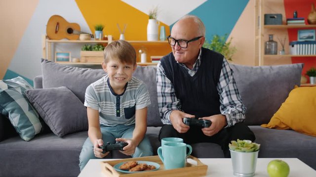 Grandfather and grandson are playing video game sitting on sofa at home using joysticks enjoying entertainment. Family and modern technology concept.