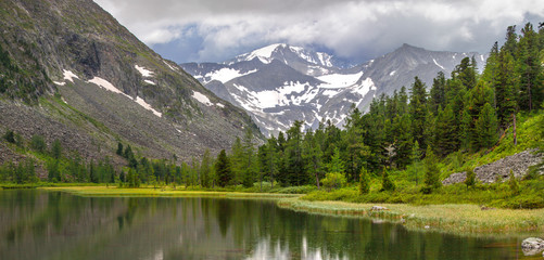 Cloudy weather, panoramic mountain landscape. Lake, forest and snow-capped peaks.
