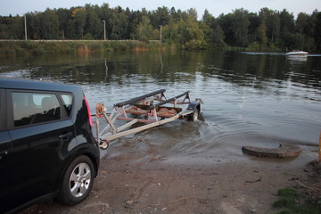 Fototapeta na wymiar Boat launch, a car with empty ship trailer in water on slip way at summer evening on floating motor boat and trees background