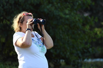 A large Caucasian woman with binocular looks from left to right on a summer day against a blurry background of green trees with copy space place