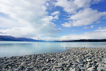 The sandy beach by the lake, behind the mountains, there are rocks and the sky.