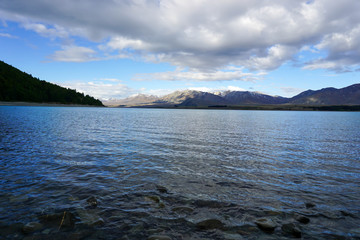 The sandy beach by the lake, behind the mountains, there are rocks and the sky.