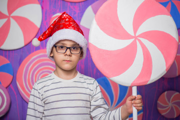 A boy in a red Christmas hat and glasses holding a toy candy in his hands, against the background of multi-colored candies
