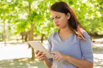 Fototapeta premium Thoughtful young woman using tablet in park. Serious lady touching screen of tablet during sunny windy day. Technology concept