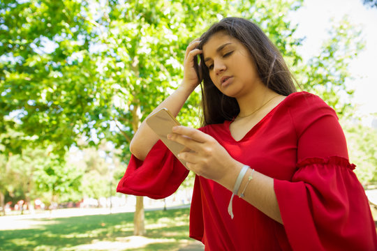 Low angle shot of confused teenage girl holding smartphone. Thoughtful teengaer looking at smartphone and rubbing head in park. Technology concept