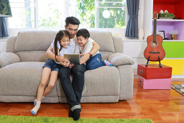happy asian dad teaching his daughter to use or play tablet on grey sofa in living room with laughing and smiling face (lifestyle with technology concept)