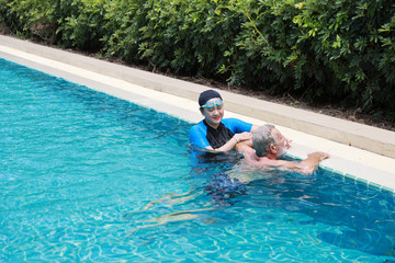 happy elderly caucasian husband and elderly asian wife swimming in pool during retirement holiday with relaxation on sunny day