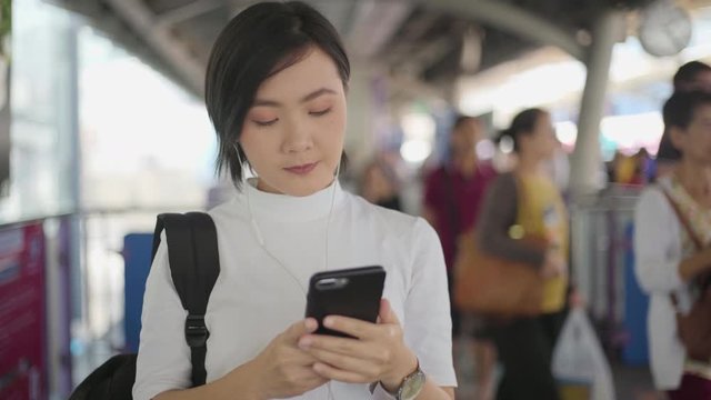 Portrait of asian woman with earphone listening music and using smartphone for chatting with friends or browsing while waiting for a train. Technology in everyday life and travel.