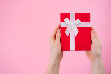 Woman hands holding red gift box wrapped with white ribbon over pink background. St. Vaventine`s Day or birthday concept.