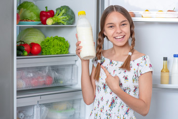 Beautiful young teen girl holding bottle of milk while standing near open fridge in kitchen at home. Portrait of pretty child choosing food in refrigerator full of healthy products.