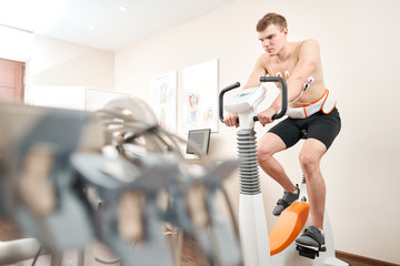 Man patient, pedaling on a bicycle ergometer stress test system for the function of heart checked. Athlete does a cardiac stress test in a medical study, monitored by the doctor. - obrazy, fototapety, plakaty
