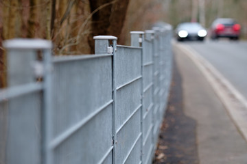 Long grey rod mat fence made of steel beside the pavement and a street with cars passing along. Seen in Germany on a moody winter day.