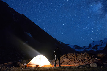 Starry night mountain landscape with glowing tent and looking at the sky person.